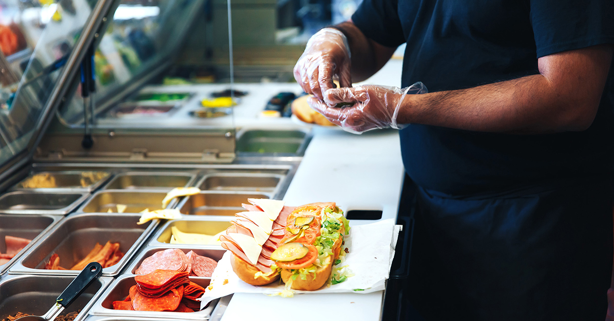 A man handling food behind a sandwich counter with plastic gloves