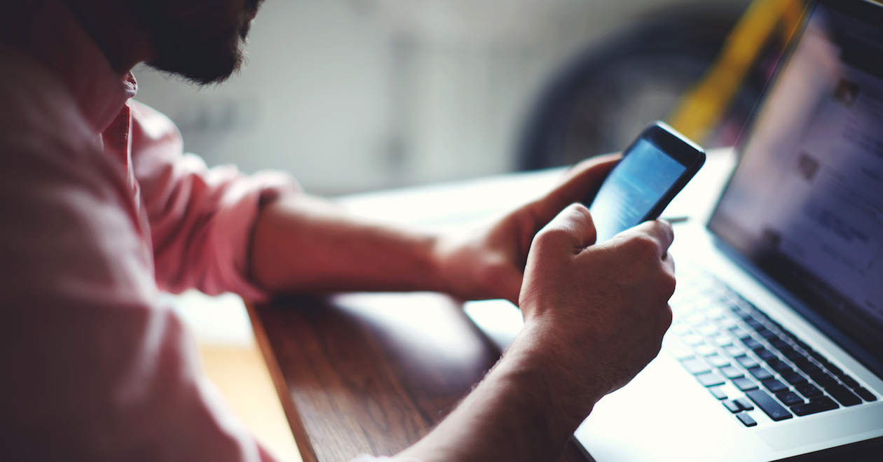 A man using a smartphone with a laptop in front of him.