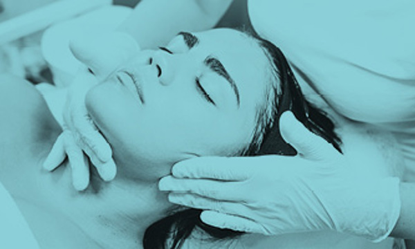 A person laying down with their eyes closed receiving a beauty treatment on their face from someone wearing cosmetic gloves.