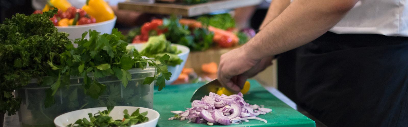 A chef's hands chopping a red onion on a green chopping board, against background of busy restaurant kitchen