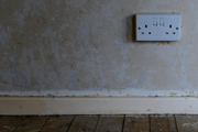 A UK double plug wall socket, showing the power supply on an unfinished interior wall with pine floor boards and staining remnants.