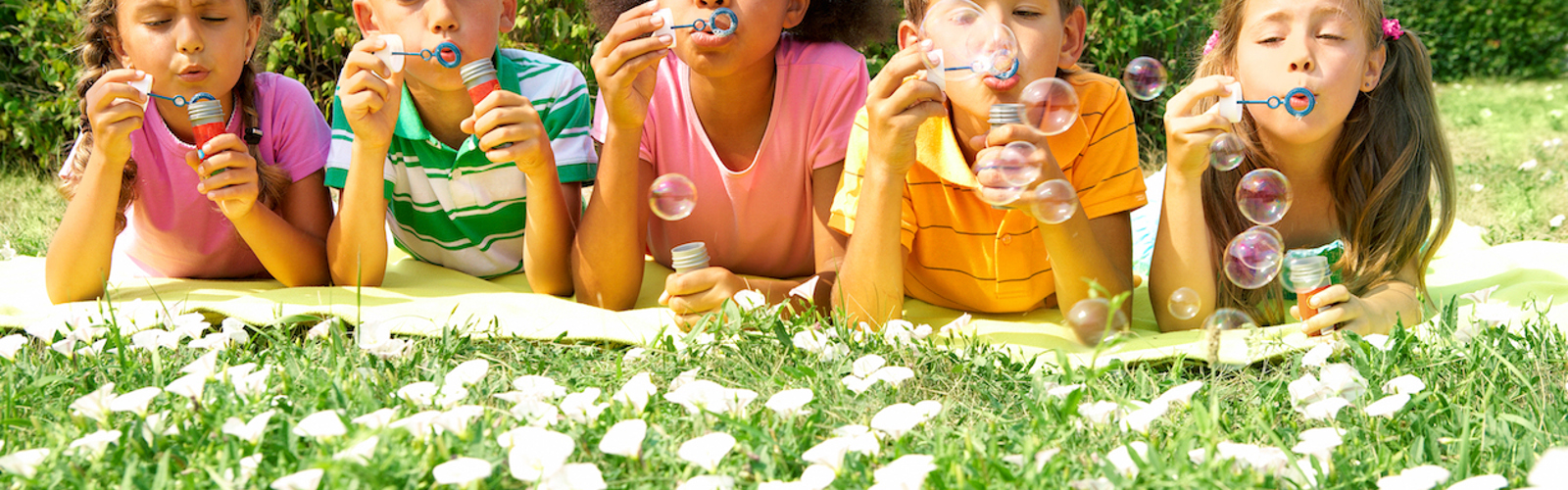 Five children lying down on a lawn full of daisies, facing camera, blowing bubbles