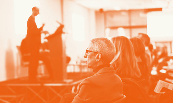 Front row audience at a conference listening to speaker stood at a podium