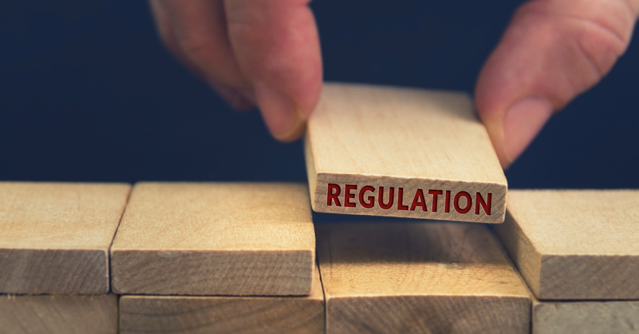 Hand placing a wooden block which reads 'Regulation' into a set of building blocks