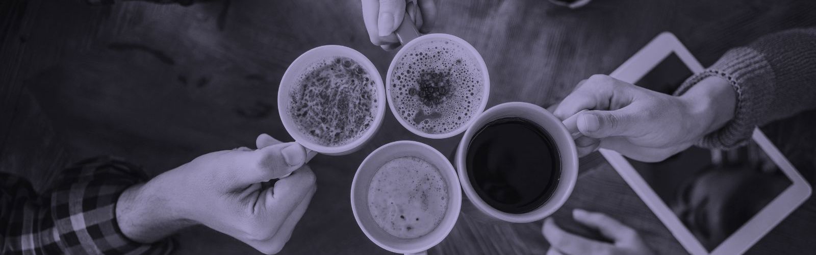 Ariel view of 4 people clinking full coffee cups with an ipad on the desk